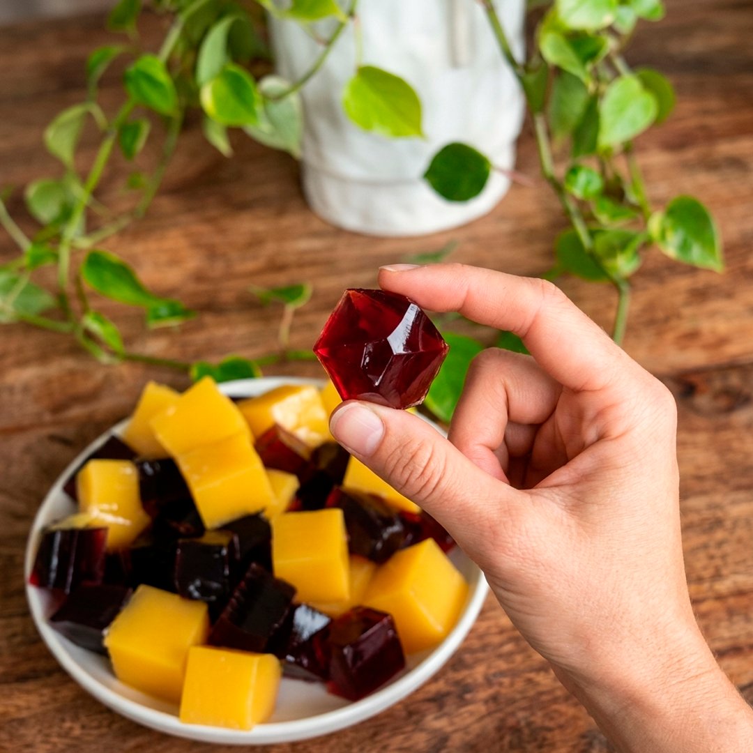 Deep crimson and golden jelly cubes on a white plate, hand holding one up to show its gem-like translucency